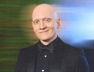 Anthony Carrigan wearing a black suit and smirking at the camera