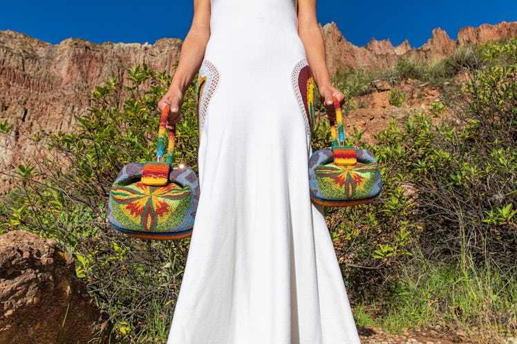 A model wearing a long white sleeveless dress while holding two multi-colored Gabriela Hearst bags. 