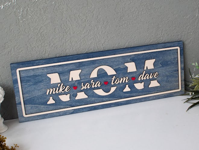 This wooden decorative sign says "Mom" across it in white lettering and has children's names across ...