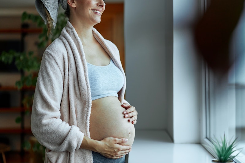 Attractive pregnant woman standing in the living room, looking out the window and holding her belly....