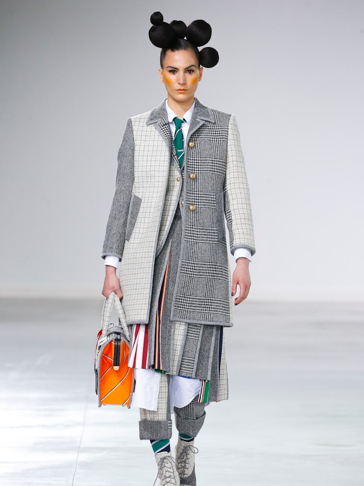 A model walking the Thom Browne Fall 2022 runway in a long grey and white jacket and orange bag 