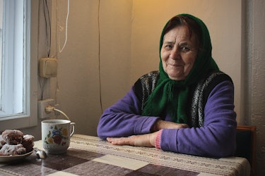 An older woman with a scarf around her head sits at a table by the window. Ukraine.