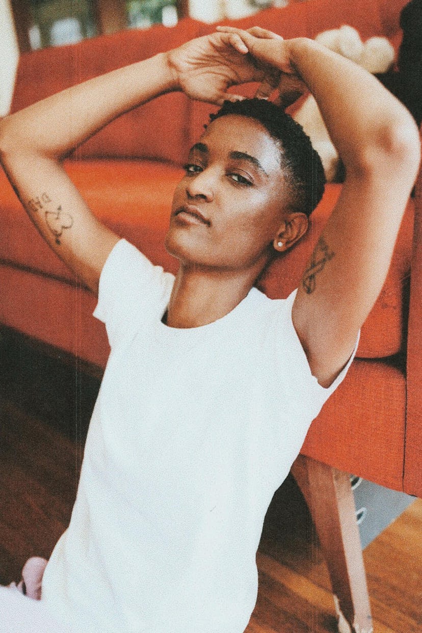 Syd leaning against an orange couch in a plain white tee 