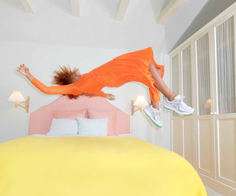 Model jumps on a bed wearing an orange dress and Ganni and New Balance's Y2K Sneaker.