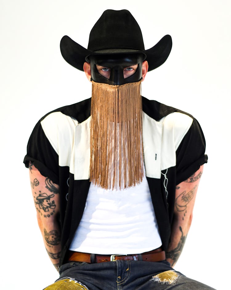 A portrait photo of Orville Peck against a white background. He wears a black hat and black mask wit...