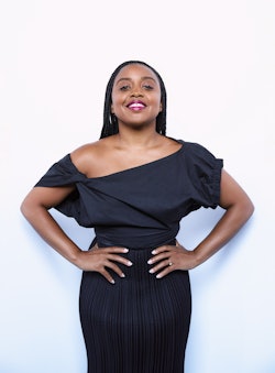 Quinta Brunson stands akimbo, smiling with her chin up in an off-the-shoulder-black dress.