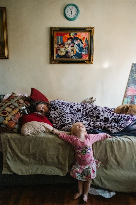 A woman laying down, covered with a blanket, sleeping, and a little girl in front of her bed, with h...