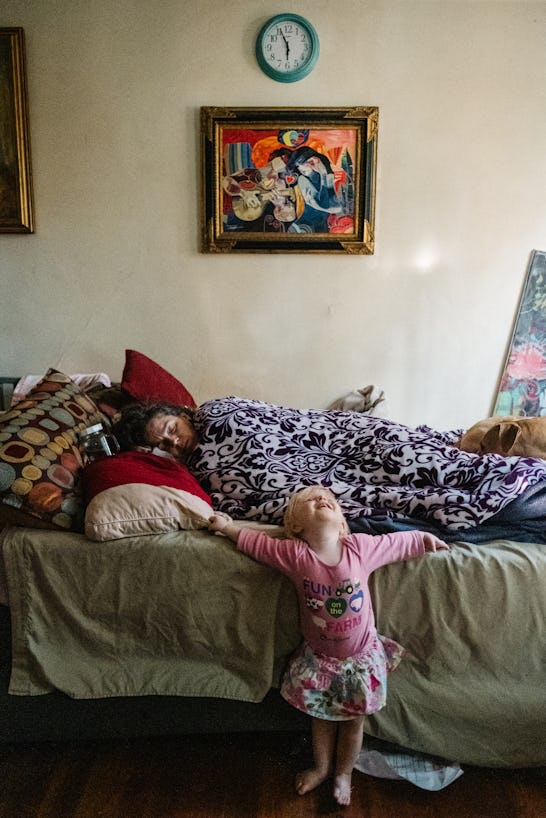 A woman laying down, covered with a blanket, sleeping, and a little girl in front of her bed, with h...