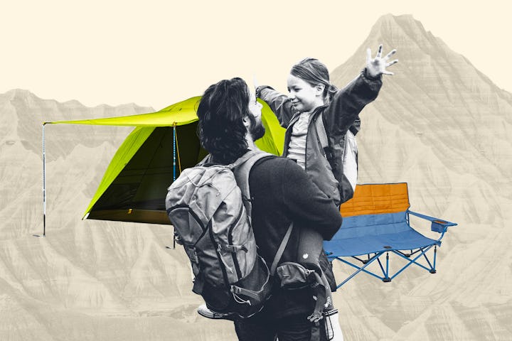A dad in hiking gear carries his daughter while there is a tent a camping chair and a mountain in fr...