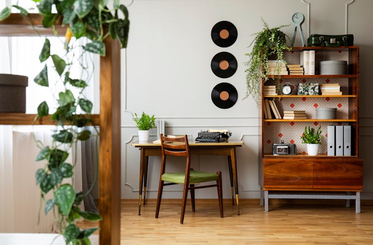 room with a shelf filled with plants on the left, a desk in the middle with three vinyl records abov...