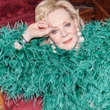 Jean Smart in a green feathered Valentino coat and Rebecca De Ravenel earrings, lounging on the red ...