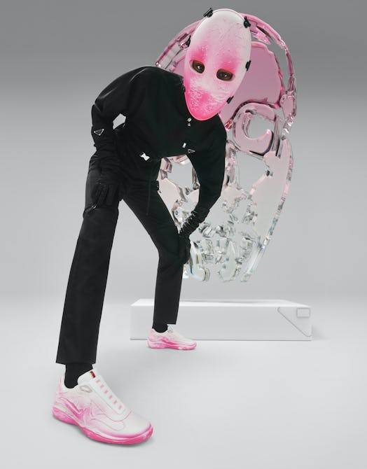 Model in a black jumpsuit wearing a pink mask covering their entire face, with pink Prada sneakers. 