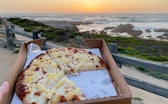 A small box of pizza held up in front of a beach sunset.