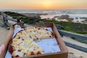 A small box of pizza held up in front of a beach sunset.