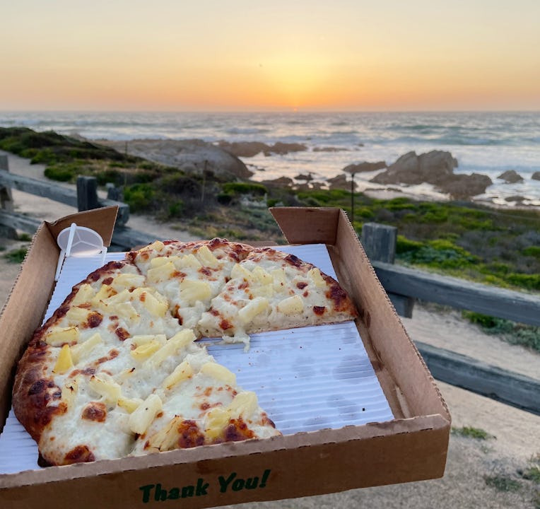 A small box of pizza held up in front of a beach sunset.