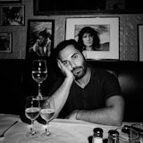 The chef and writer Andy Baraghani sitting at a restaurant table with his hand to his cheek