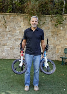 Actor George Clooney standing in a walled garden holding two children's bicycles