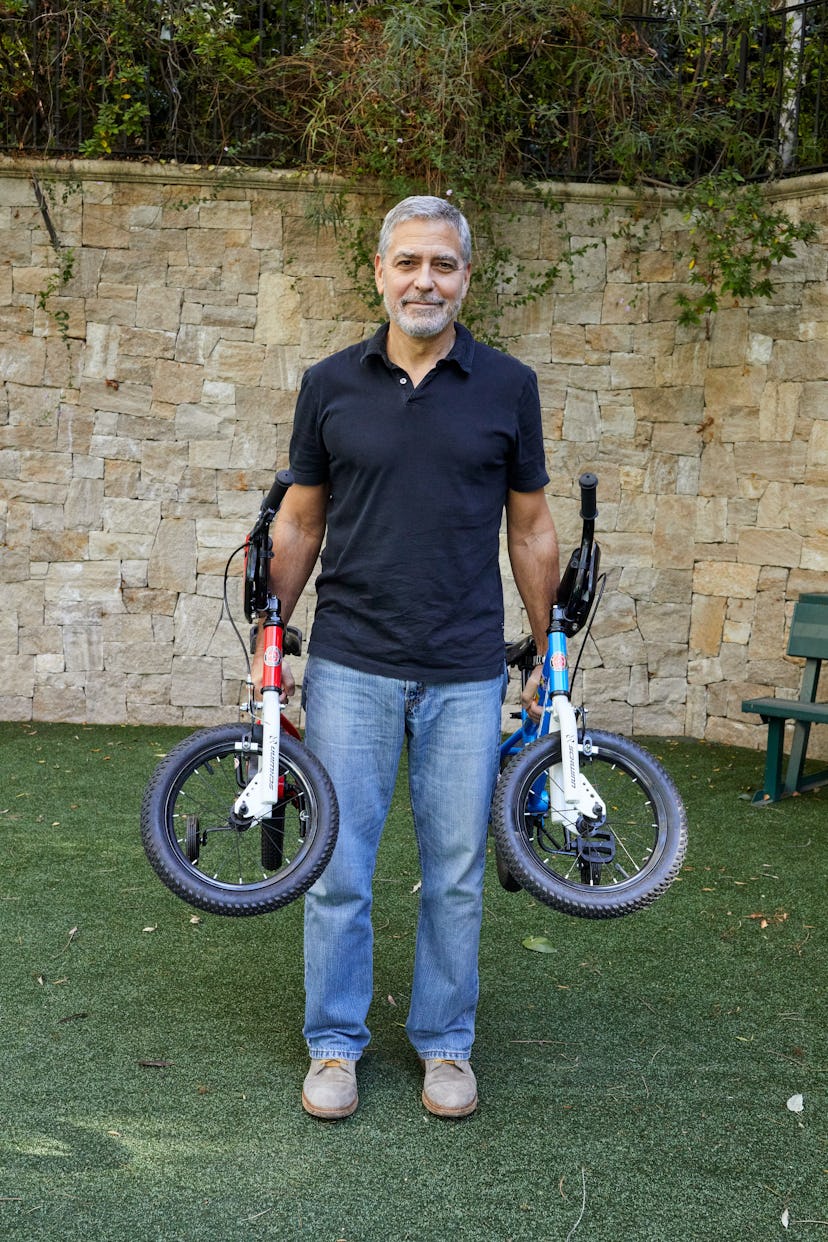 Actor George Clooney standing in a walled garden holding two children's bicycles