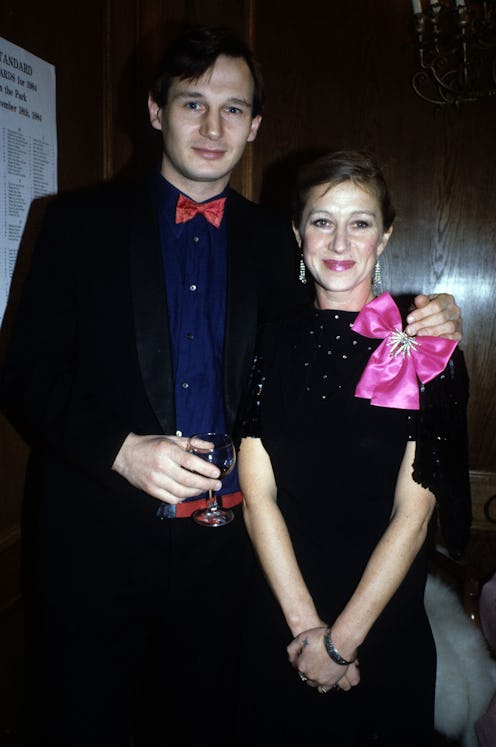 A young liam neeson and helen mirren pose side by side. He's wearing a black suit with a red shirt, ...