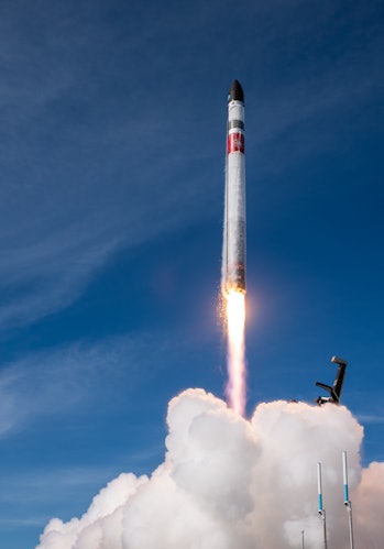 A white rocket launches into the sky, and a big white plume is visible underneath it. The sky is a d...