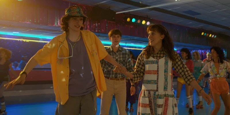 mike wheeler and eleven hold hands at a skate rink wearing '80s clothes in stranger things season 4