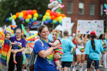A parent holds their baby, dressed in rainbow, at a Pride parade.