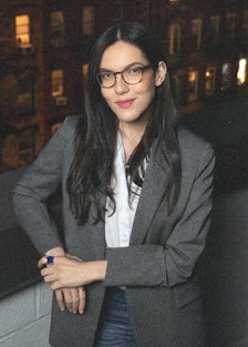 Sloane Crosley wearing glasses and a grey blazer, city in the background behind her