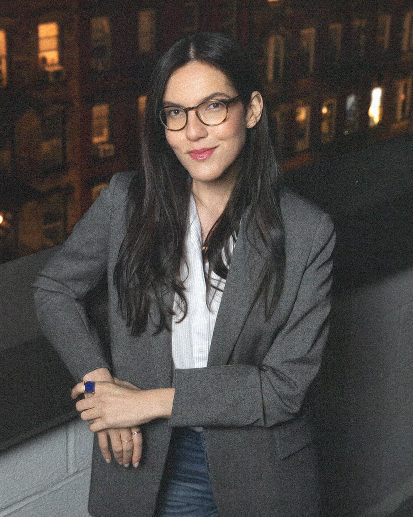 Sloane Crosley wearing glasses and a grey blazer, city in the background behind her