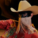 A portrait of Orville Peck wearing a beige cowboy hat, black mask with gold fringe, and a rust color...