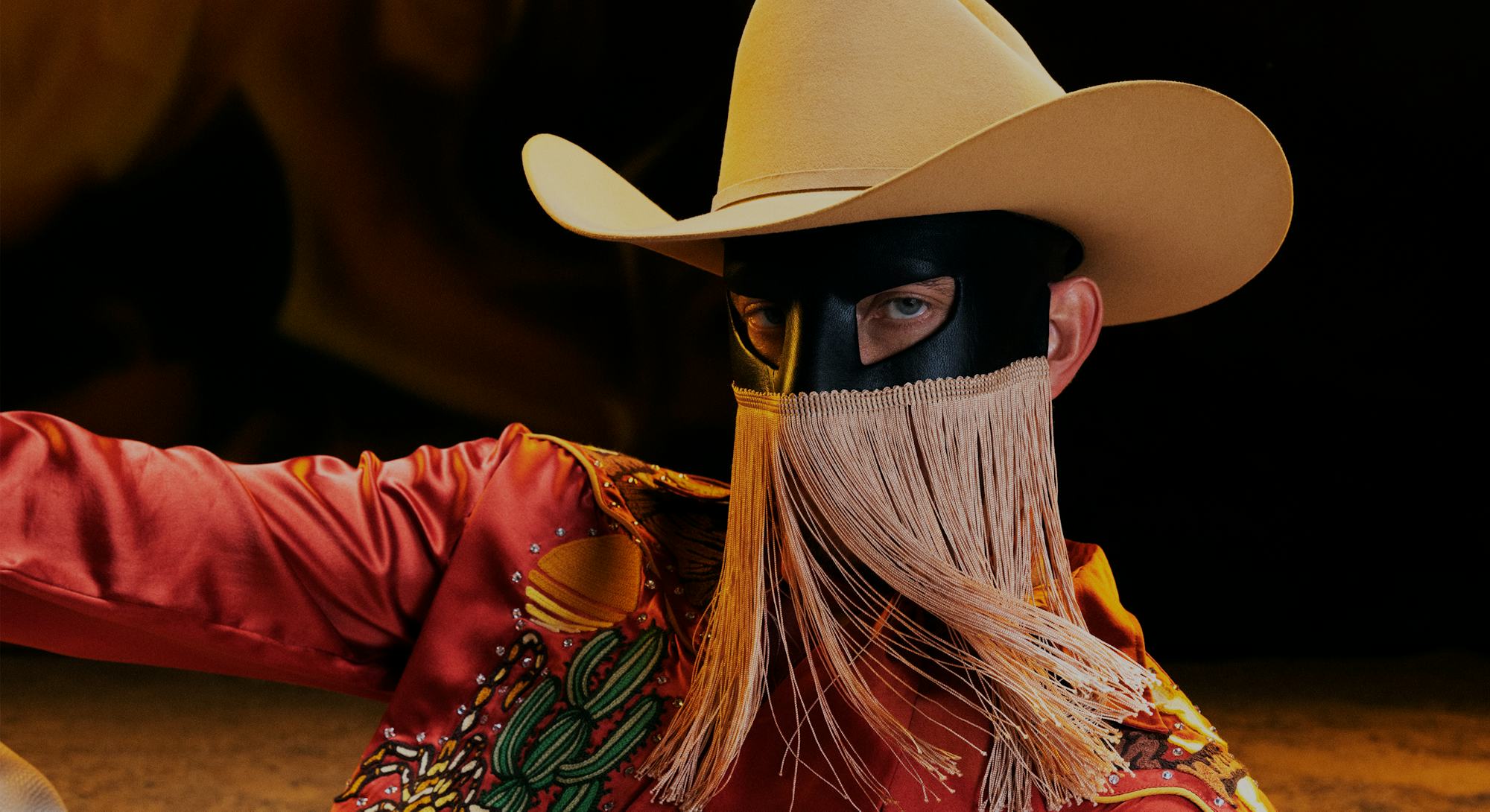 A portrait of Orville Peck wearing a beige cowboy hat, black mask with gold fringe, and a rust color...