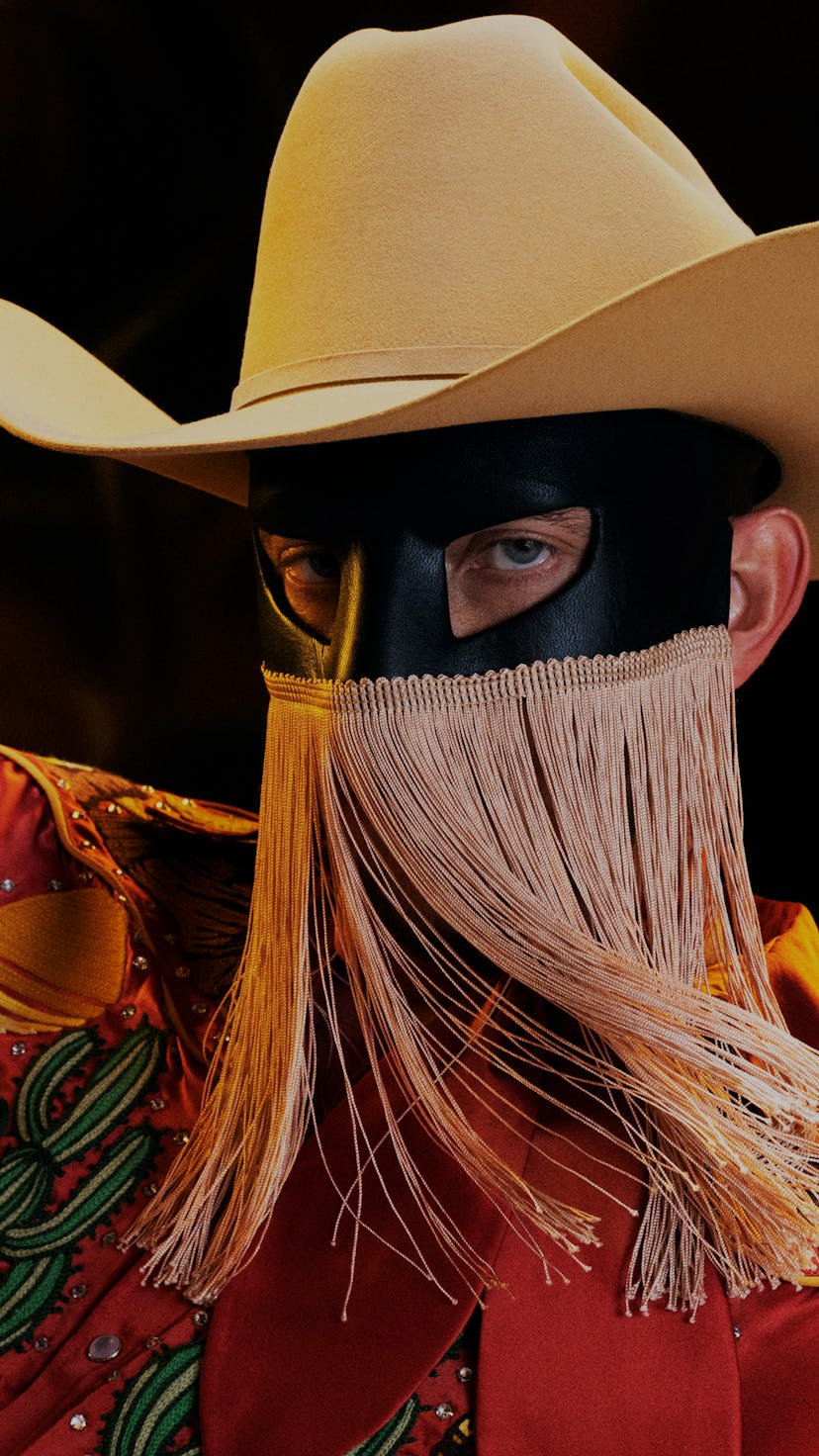 A portrait of Orville Peck wearing a beige cowboy hat, black mask with gold fringe, and a rust color...