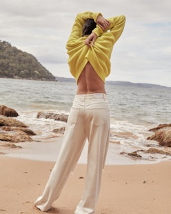 A woman in white denim pants taking off a yellow sweater while standing on a beach