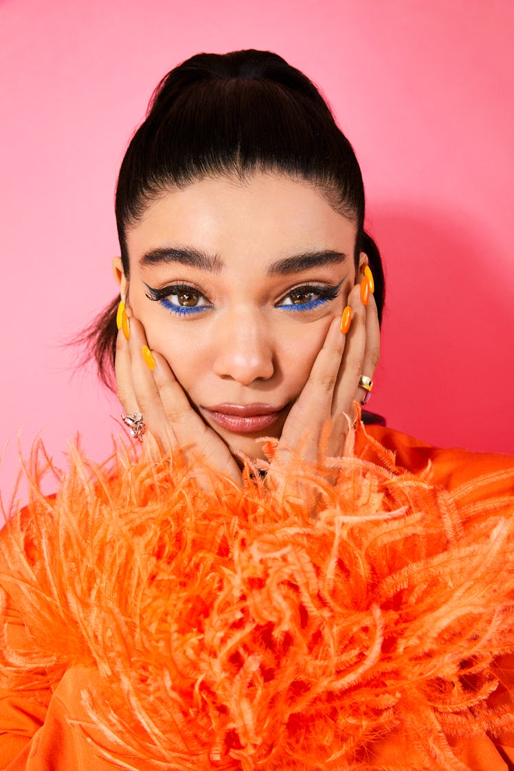 Actress Iman Vellani posing in a bright orange outfit while rocking a blue eyeshadow