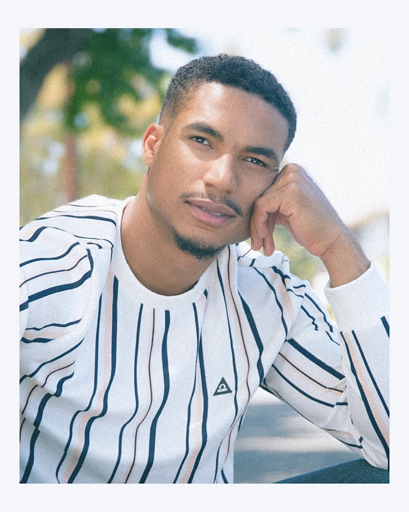 A portrait of Greg Tarzan sitting on a curb, wearing a white striped shirt, with his hand on his che...