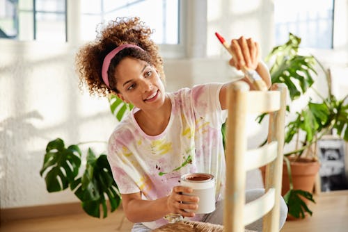 Smiling woman in messy casuals applying paint on chair. Young female is holding paint can. She is up...