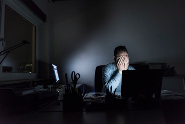 A depressed man in a dark office rests his face in his hands.