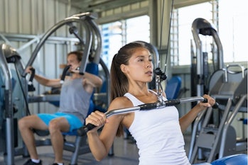 A woman in the gym performs a lat pulldown. A man behind her is using a machine to perform a chest p...