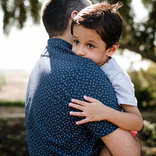 An autistic boy hugs his dad as they stand under a tree.