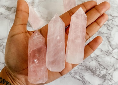 Close up of a palm holding three rose quartz crystal towers on a white marble background, a heart ch...