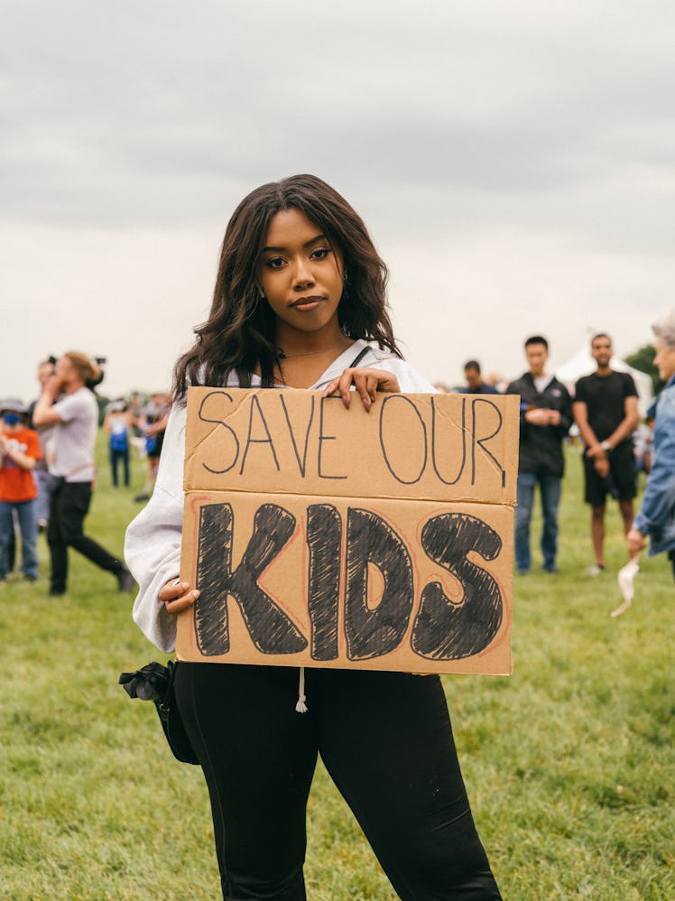 A protestor stands with a homemade sign that reads, "SAVE OUR KIDS". Other attendees stand in the di...