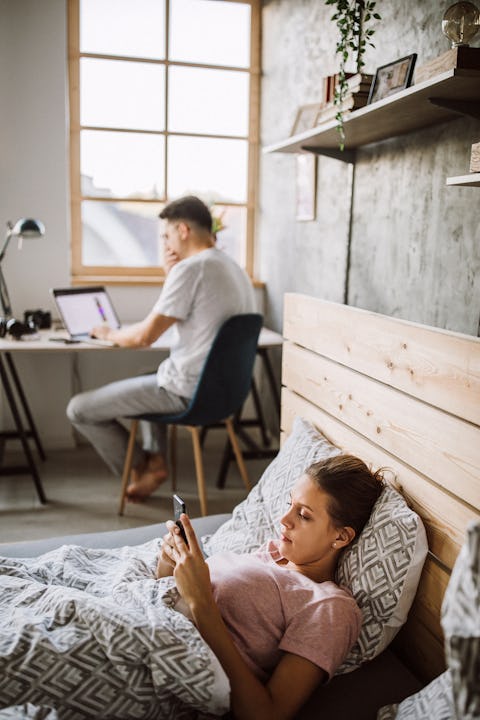 A woman laying on a bed checking her phone while a man is sitting at his desk with a laptop