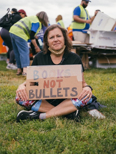 Protestor sitting cross-legged on the grass, holding a sign in her lap that reads, "BOOKS NOT BULLET...