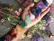 Woman's hand holding a line up of eleven different colored crystals for the chakras above a spread o...