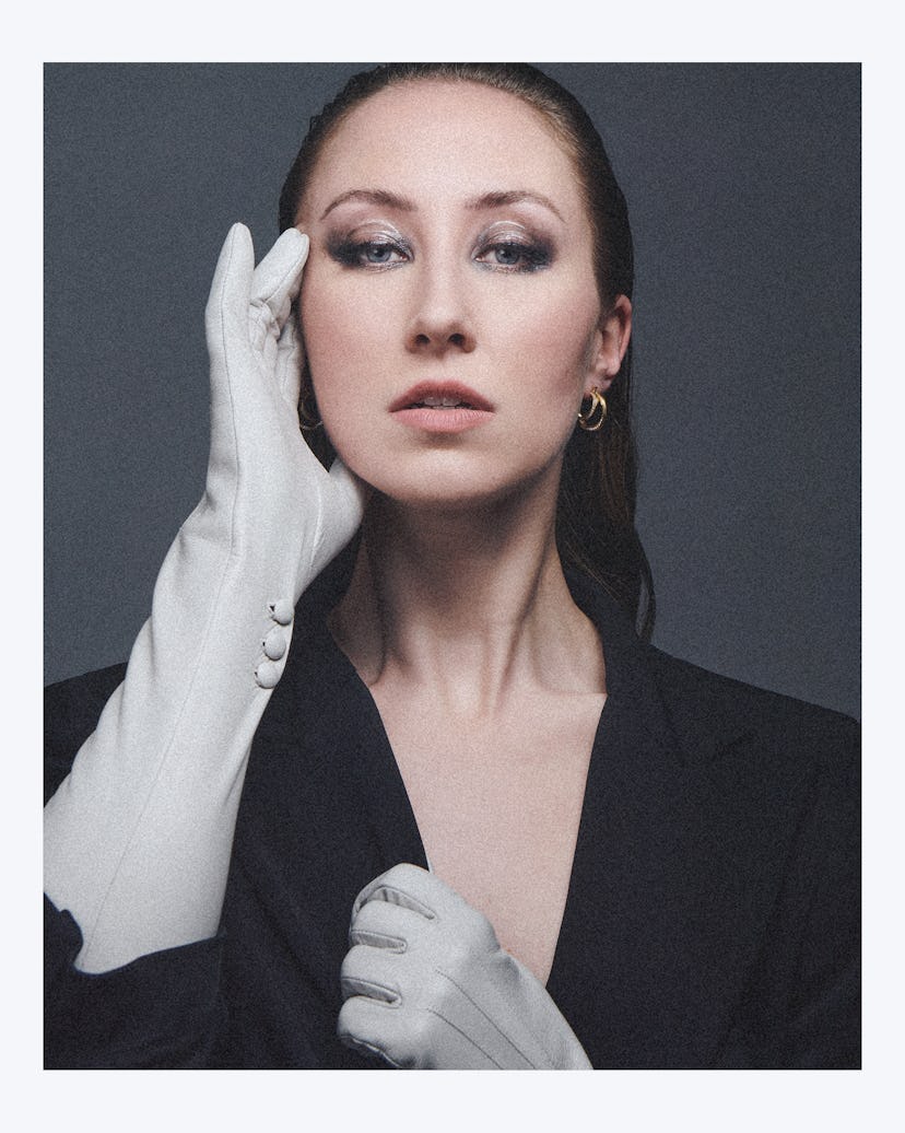 A portrait of Erin Doherty, wearing a black suit and white glove, with her hand to her face