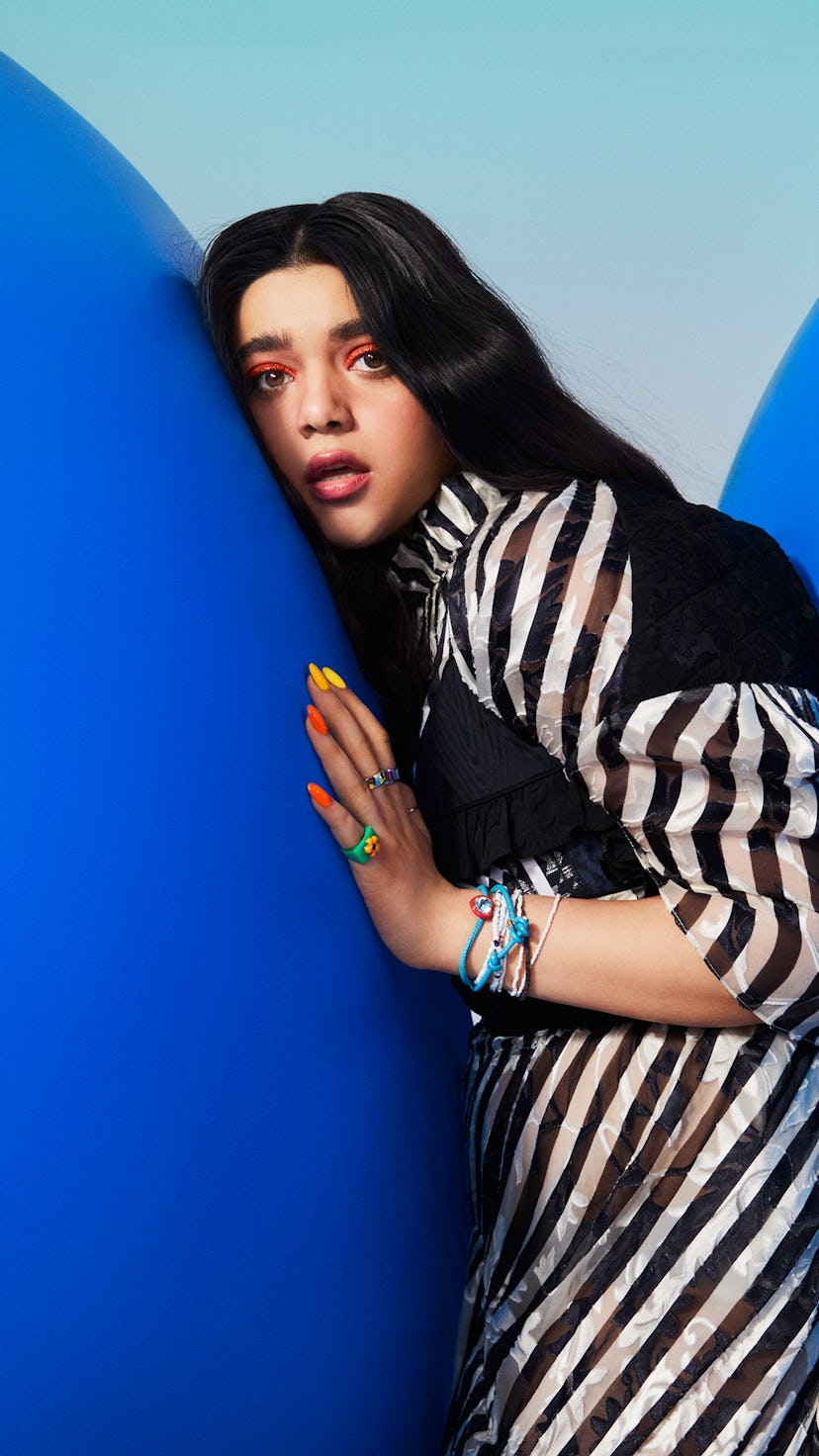 Actress Iman Vellani posing on a blue background wearing black and white outfit with bright orange m...