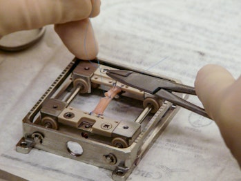 Two gloved hands work on a tiny metal frame. At the frame's center is a pale pink band, which is act...