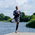 A man stands in tree pose on a dock surrounded by water, with trees in the distance.