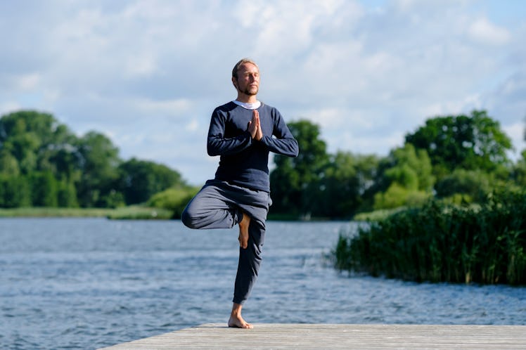 A man stands in tree pose on a dock surrounded by water, with trees in the distance.