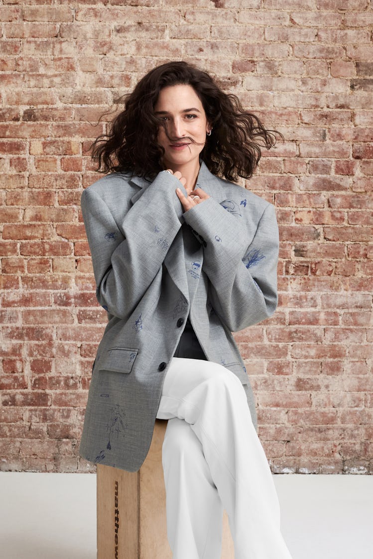 Jenny Slate in a large Vetements jacket sitting cutely before a brick wall