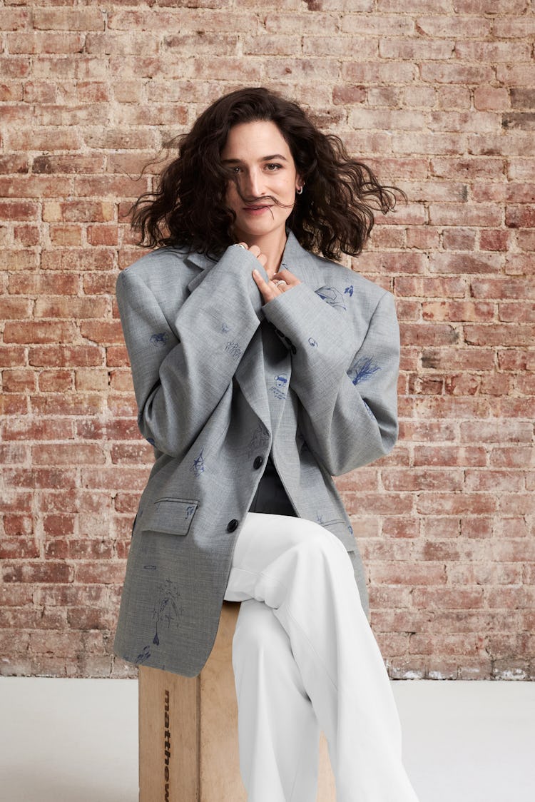 Jenny Slate in a large Vetements jacket sitting cutely before a brick wall